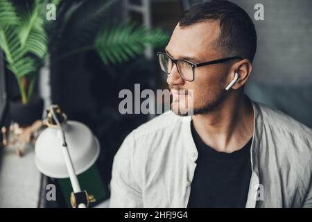 Porträt eines ernsthaften kaukasischen Mannes in einer Brille, der mit kabellosen Kopfhörern wegschaut. Moderner Stil. Stockfoto