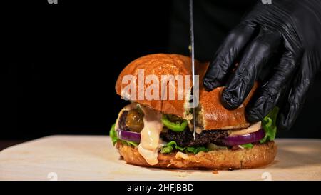 Schneiden Handwerk Burger ist Kochen auf schwarzem Hintergrund. Bestehen salsa Sauce, Salat, rote Zwiebel, Gurke, Käse, Chili, Paprika, Brötchen, marmoriertes Fleisch Rindfleisch. Stockfoto