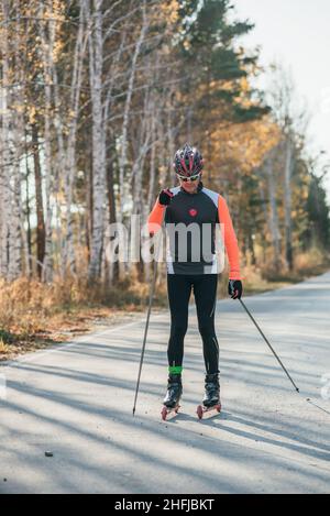 Training ein Athlet auf der Walze Skater. Biathlon Fahrt auf dem Roller Ski mit Skistöcke, im Helm. Herbst Training. Roller Sport. Stockfoto