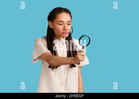 Attraktive neugierige Frau mit schwarzen Dreadlocks, die durch die Lupe wegschaut, ausspioniert, etwas findet, den Tatort erkundet, inspiziert. Innenaufnahme des Studios isoliert auf blauem Hintergrund. Stockfoto