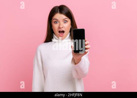 Staunende Brünette weibliche zeigt Handy mit leerem Display mit Kopieplatz für Werbung, trägt weißen lässigen Stil Pullover. Innenaufnahme des Studios isoliert auf rosa Hintergrund. Stockfoto