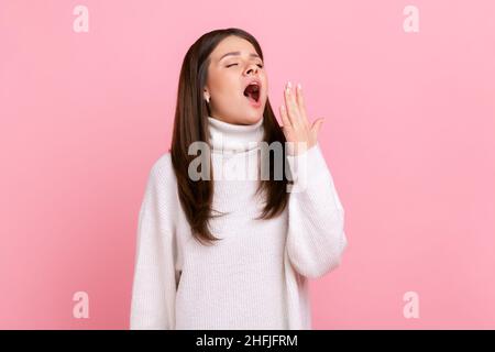 Verschlammte junge Frau, die mit geschlossenen Augen gähnend steht und den Mund bedeckt, sich erschöpft fühlt und sich ausruhen muss, trägt einen weißen Pullover im lässigen Stil. Innenaufnahme des Studios isoliert auf rosa Hintergrund. Stockfoto