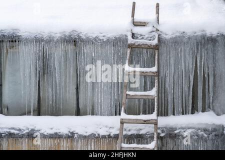 Alte Holzleiter, die an der Wand gefriert und mit gefrorenem Eis und Eiszapfen bedeckt ist. Winterliches kaltes Klima Stockfoto