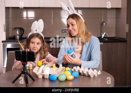 kaukasische Mutter und Tochter in einem Verband auf den Ohren eines Kaninchens sitzen in der Küche zu Hause. Dekoration Ostereier mit Farbe Blogger Broa Stockfoto