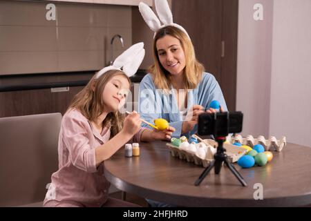 kaukasische Mutter und Tochter in einem Verband auf den Ohren eines Kaninchens sitzen in der Küche zu Hause. Dekoration Ostereier mit Farbe Blogger Broa Stockfoto