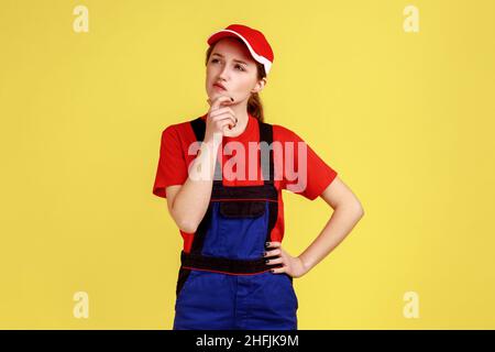 Porträt einer nachdenklichen Frau, die das Kinn hält, mit nachdenklichem Gesichtsausdruck wegschaut, Overalls und roter Mütze trägt. Innenaufnahme des Studios isoliert auf gelbem Hintergrund. Stockfoto