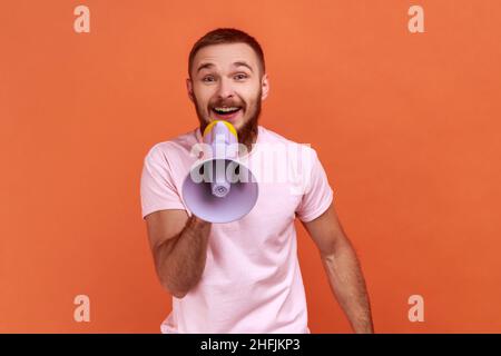 Porträt eines optimistischen bärtigen Mannes, der im Megaphon schreit und wichtige Informationen mit positivem Ton ankündigt, die Kamera anschaut und ein pinkes T-Shirt trägt. Innenaufnahme des Studios isoliert auf orangefarbenem Hintergrund. Stockfoto