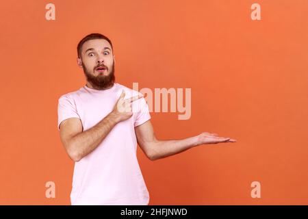 Porträt eines überraschten bärtigen Mannes, der mit dem Finger auf den leeren Kopierraum zeigt, mit Erstaunen, Platz für Werbung, in rosa T-Shirt. Innenaufnahme des Studios isoliert auf orangefarbenem Hintergrund. Stockfoto