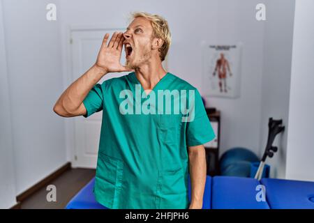 Junger blonder Mann in Physiotherapeut-Uniform steht in der Klinik und schreit laut zur Seite mit der Hand auf dem Mund. Kommunikationskonzept. Stockfoto