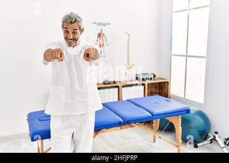 hispanischer Therapeut im mittleren Alter, der in der Schmerzklinik arbeitet, zeigt auf Sie und die Kamera mit den Fingern, lächelt positiv und fröhlich Stockfoto