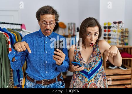 Interrassisches Paar im mittleren Alter im Einzelhandelsgeschäft mit Smartphone, das nach unten zeigte, mit Fingern, die Werbung zeigten, überraschte das Gesicht und den offenen Mund Stockfoto