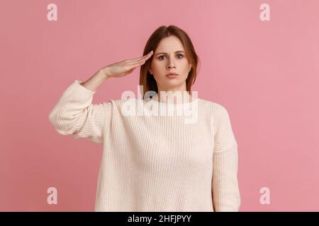 Porträt einer verantwortungsbewussten jungen erwachsenen blonden Frau, die den Kommandanten grüßend, gehorsam und mit weißem Pullover anhört. Innenaufnahme des Studios isoliert auf rosa Hintergrund. Stockfoto