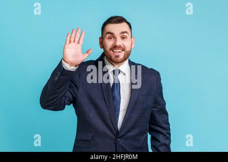 Hallo. Portrait von freundlichen froh gut aussehenden Brünette Mann mit Bart tragen offiziellen Stil Anzug zeigt hallo Geste mit winkender Hand und lächelnd aufrichtig Indoor Studio Schuss auf blauem Hintergrund isoliert. Stockfoto