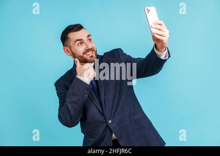 Porträt eines freundlichen, gutaussehenden Mannes, der einen Anzug im offiziellen Stil trägt und den Daumen nach oben zeigt und lächelnd auf die Kamera seines Smartphones blickt und dabei bloggt. Innenaufnahme des Studios isoliert auf blauem Hintergrund. Stockfoto