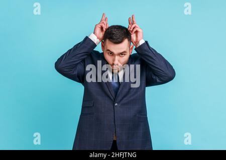 Aggressiver Schläger mit Bart und offiziellem Anzug, der die Finger über dem Kopf hält und Hörner zeigt, arrogant und stur, angriffbereit. Innenaufnahme des Studios isoliert auf blauem Hintergrund. Stockfoto