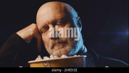 Ein älterer Mann, der einen langweiligen Film im Kino sieht und Popcorn isst, schläft mit geschlossenen Augen Stockfoto