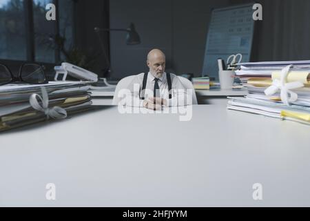 Kleiner Geschäftsmann, der am Schreibtisch sitzt und sich die Stapel von Papierkram, überwältigende Arbeit und Verantwortlichkeiten anschaut Stockfoto