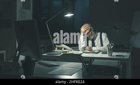 Gestresster Geschäftsführer, der am Schreibtisch sitzt und nachts Überstunden macht, ist überfordert und müde Stockfoto