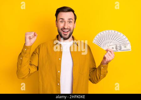 Foto von lustigen glücklichen jungen Kerl tragen lässige Kleidung lächelnd steigende Faust hält Geld Stapel isoliert gelbe Farbe Hintergrund Stockfoto