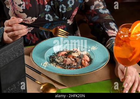 Weibliche Hände halten Glas Cocktail und Gabel gehen Tintenfisch Tinte Pasta mit Meeresfrüchten essen Stockfoto
