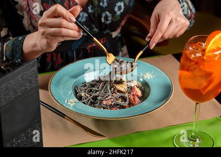 Eine kurze Aufnahme einer Frau, die schwarze Pasta mit Meeresfrüchten und Orangenspritz isst Stockfoto