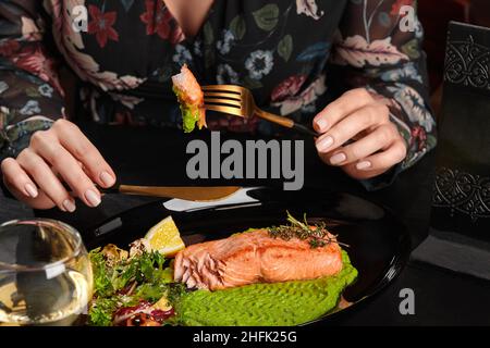 Weibliche Hände halten Messer und Gabel mit gebackenen Forellen Steak Stück serviert mit pürierten Erbsen, Salat, Zitrone Stockfoto