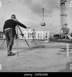 Craigshill, Livingston, West Lothian, Schottland, 01/10/1965. Zwei Vermesser bei der Arbeit auf einem Jespersen-Baugelände, wahrscheinlich in der Craigshill-Entwicklung in Livingston, mit einem Kran im Hintergrund, der eine Palette von einem Anhänger auslädt. Im Jahr 1963 kauften John Laing und Son Ltd die Rechte an dem dänischen industrialisierten Bausystem Jespersen (manchmal auch als Jesperson bezeichnet). Das Unternehmen baute Fabriken in Schottland, Hampshire und Lancashire, die Fertigteile und Betonfertigteile von Jespersen produzierten, wodurch der Wohnungsbau rationalisiert werden konnte, was Zeit und Geld einspar Stockfoto