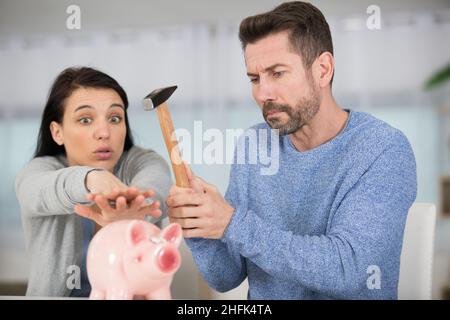 Frau, die das Sparschwein davor schützt, vom Ehemann mit Hammer zerschlagen zu werden Stockfoto