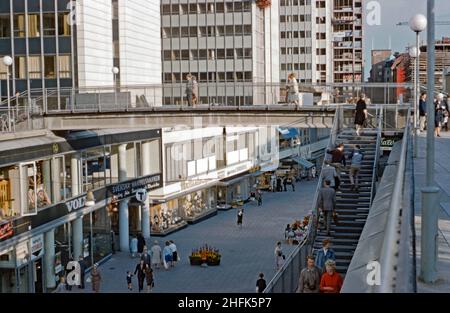 Blick auf die Fußgängerzone Sergelgatan unter den Hötorget-Gebäuden (links) im zentralen Norrmalm-Viertel, Stockholm, Schweden im Jahr 1960s. Die Fußgängerbrücke über das Einkaufszentrum scheint seitdem entfernt worden zu sein. Im Hintergrund werden neue Gebäude errichtet. Die Hötorget-Gebäude sind fünf moderne Bürohochhäuser. Dieses Bild stammt von einer Amateur-35mm-Farbtransparenz – einem Vintage-1960s-Foto. Stockfoto