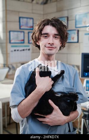 Porträt eines jungen männlichen Tierarztes in Uniform, der eine schwarze Katze in den Händen hält und die Kamera in der Tierarztklinik sieht Stockfoto