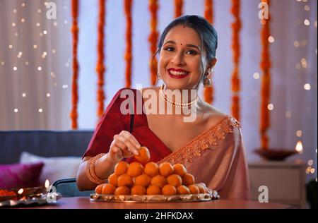 Eine wunderschöne Frau, die einen Laddoo in den Teller steckt und mit diwali-Dekoration und Lichtern lächelt. Stockfoto