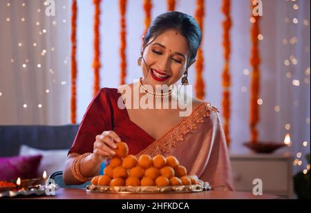 Eine wunderschöne Frau, die einen Laddoo in den Teller steckt und mit diwali-Dekoration und Lichtern lächelt. Stockfoto