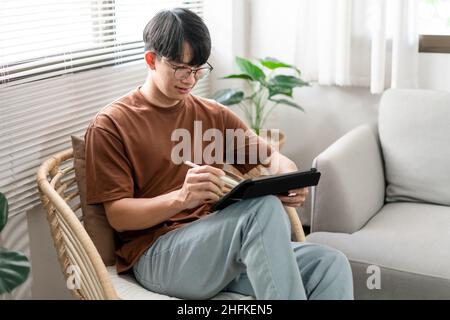 Technologiekonzept der junge Mann, der auf dem Stuhl sitzt und sein iPad hält und etwas darauf notiert. Stockfoto