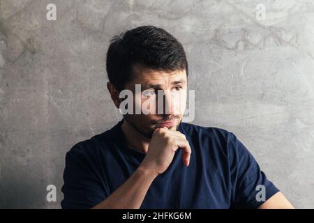 Schöner nachdenklicher, ernsthafter bärtiger Mann mit der Hand am Kinn, der im modernen Büro gegen eine Betonwand sitzt Stockfoto