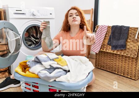 Junge Rothaarige Frau, die schmutzige Wäsche in die Waschmaschine legt, zeigt mit dem Finger nach oben und wütend und zeigt keine Geste Stockfoto