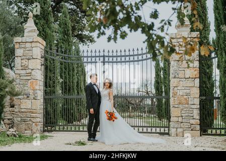 Fröhliches, stilvolles und lächelndes Paar, das am Hochzeitstag in der Toskana, Italien, unterwegs ist. Die Braut und der Bräutigam gehen die Straße entlang durch die Hände. Ein stylischer Junge Stockfoto