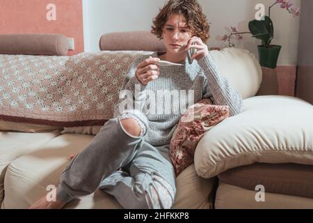 Ziemlich krankes Teenager-Mädchen mit Thermometer reden auf dem Smartphone mit Arzt, während auf dem Sofa zu Hause sitzen. Schöne ungesunde Teenager-Mädchen Stockfoto