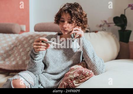 Ziemlich krankes Teenager-Mädchen mit Thermometer reden auf dem Smartphone mit Arzt, während auf dem Sofa zu Hause sitzen. Schöne ungesunde Teenager-Mädchen Stockfoto