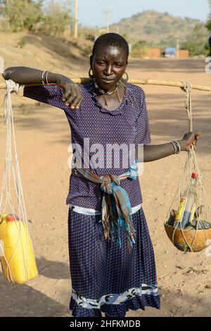 ÄTHIOPIEN, Provinz Benishangul-Gumuz, Stadtdebatte, Gumuz-Frauen, die Waren mit Joch vom Markt in ihr Dorf transportieren, Flasche Gin / AETHIOPIEN, Provinz Benishangul-Gumuz, Stadtdebatte, Gumuz Frauen mit Tragjoch kommen vom Markt, Flasche Gin in der Kalabasse Stockfoto