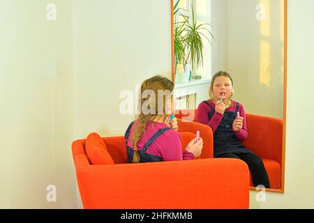 Beauty- und Modekonzept. Kind mit schönen Wangen lächelt und malt drinnen Lippen. Kleines Mädchen hält Lippenstift oder Mascara vor dem Spiegel in der Stockfoto