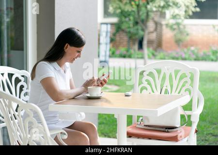 Eine junge Frau in einem weißen Kleid benutzt das Telefon, schreibt SMS oder Boten. Sommercafe auf der Straße mit einer Tasse Kaffee. Stockfoto