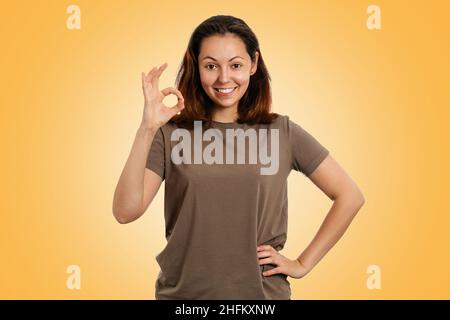 Emotionen. Das Porträt einer jungen kaukasischen Frau mit einem Lächeln zeigt die OK-Geste. Gelber Hintergrund. Menschliche Emotionen, Gesichtsausdruck Konzept. Stockfoto