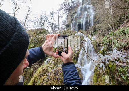 Bad Urach, Deutschland. 16th Januar 2022. Ein Mann fotografiert den Wasserfall bei Bad Urach mit seinem Handy. Der Urach-Wasserfall gehört zu den Sehenswürdigkeiten des UNESCO Global Geopark Schwäbische Alb und des UNESCO-anerkannten Biosphärenreservats Schwäbische Alb. Quelle: Thomas Warnack/dpa/Alamy Live News Stockfoto