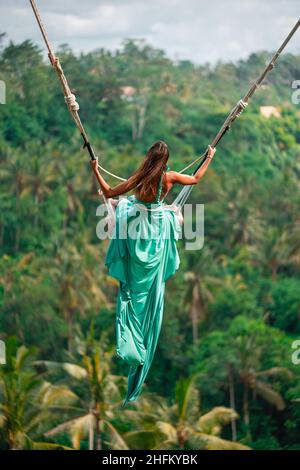Gebräunte junge Frau, die auf einer langen Schaukel reitet ist. Insel Bali. Tropischer Wald im Hintergrund. Reisen und Freude. Stockfoto