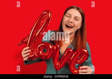 Glückliche 20s 30s Frau hält rote Folie Liebe Wort Ballons über rotem Hintergrund Stockfoto