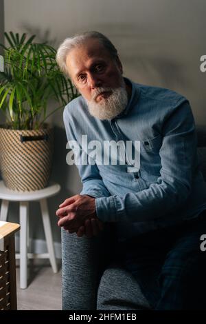 Vertikales Porträt eines selbstbewussten älteren grauhaarigen Mannes, der auf dem Sofa an der Nachttischlampe im dunklen Wohnzimmer sitzt und eine Kamera sieht. Stockfoto