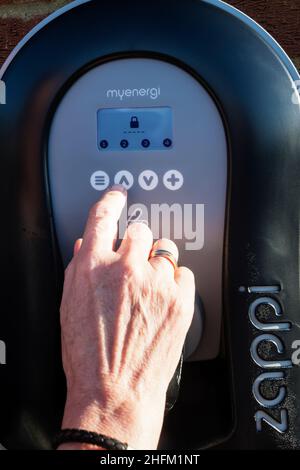 Frau, die eine PIN eingibt, um ein Ladegerät für Elektroautos von Zappi zu entsperren, das an der Außenwand einer privaten Garage befestigt ist. Sicherheitsfunktion zur Vermeidung von Stromdiebstahl. Stockfoto