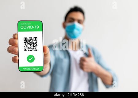 Impfpass. Glücklicher Kerl zeigt mobile App mit qr-Code auf Handy-Bildschirm, gestikuliert Daumen nach oben Stockfoto