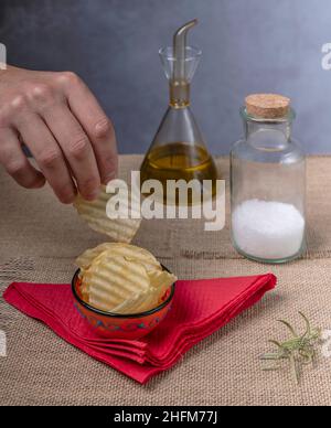 Eine männliche Hand nimmt einen Kartoffelchip aus einer orangefarbenen Untertasse mit Salz und Olivenöl in der Nähe Stockfoto