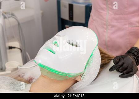 Frau liegt im Bett mit LED-Lichttherapie Gesichtsmaske und entspannen. LED-Lichtmaske für die Gesichtshautbehandlung, franfessionelles Kosmetologieverfahren. Stockfoto